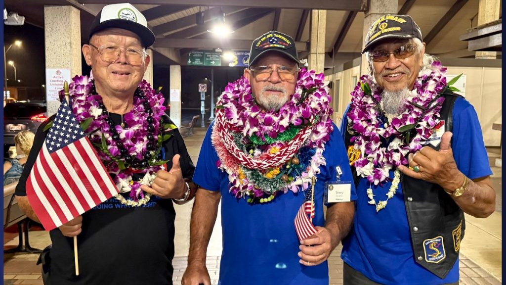 Veterans Edward Kawamura, Terry Daligdig, and Robert "Bobbie" Paik, along with community members and family, wait outside baggage claim B at Lihue Airport as the rain subsides. (Photo Credit: Xiomara Yamileth/ Kauaiʻi Now)