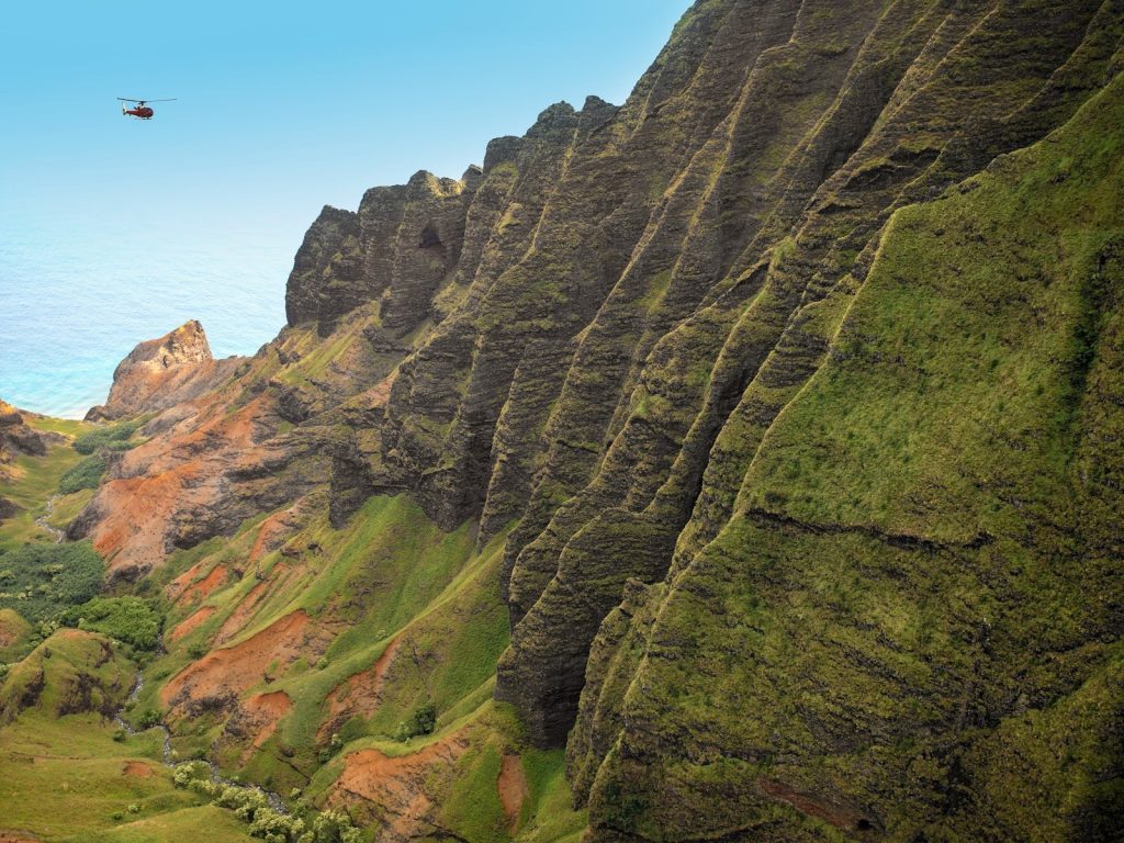 A helicopter glides past the emerald cliffs of the Nāpali Coast, where spring rains bring fresh green life to Kaua‘i’s dramatic landscape.
