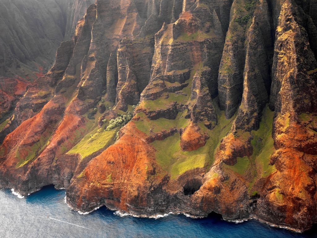 Golden summer light illuminates the rugged cliffs of the Nāpali Coast, where red and green ridges meet the deep blue Pacific.