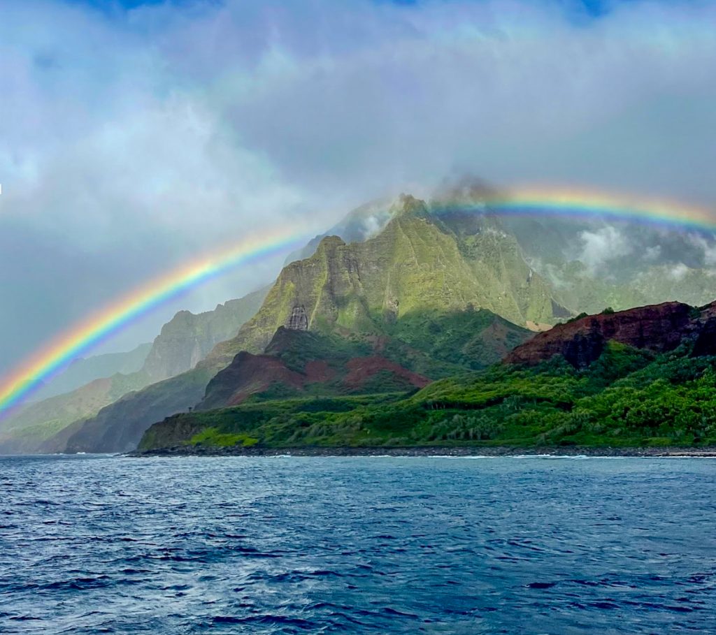 The Nāpali Coast reveals one of its most fleeting and breathtaking moments — a full arc rainbow of color stretched across emerald cliffs, viewed from the water as only a boat tour can offer. (Photo Credit: Ilana Maxwell)
