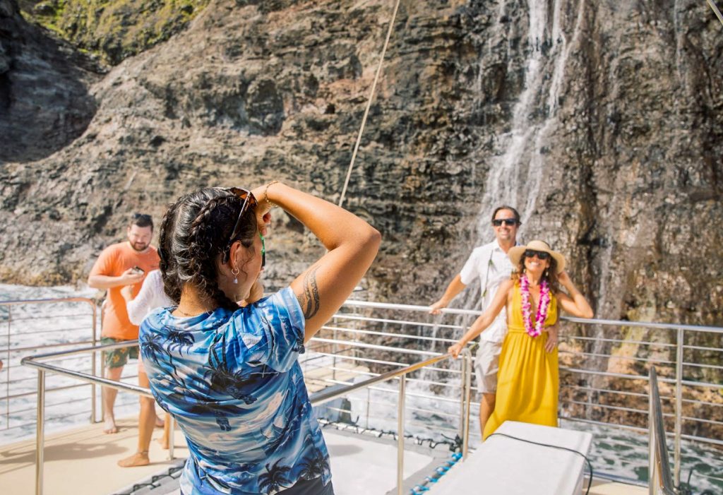 Guests aboard a Blue Dolphin Kauaʻi catamaran pause for a photo in front of one of Nāpali's ephemeral waterfalls — the kind that only appear after a good rain and disappear just as quickly. (Photo Credit: Ilana Maxwell)