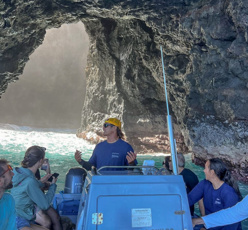 Inside the open-ceiling sea cave along the Nāpali Coast, a Blue Dolphin Kauaʻi captain shares the story of this remarkable place with guests gathered beneath ancient lava rock walls. (Photo Credit: Ilana Maxwell)